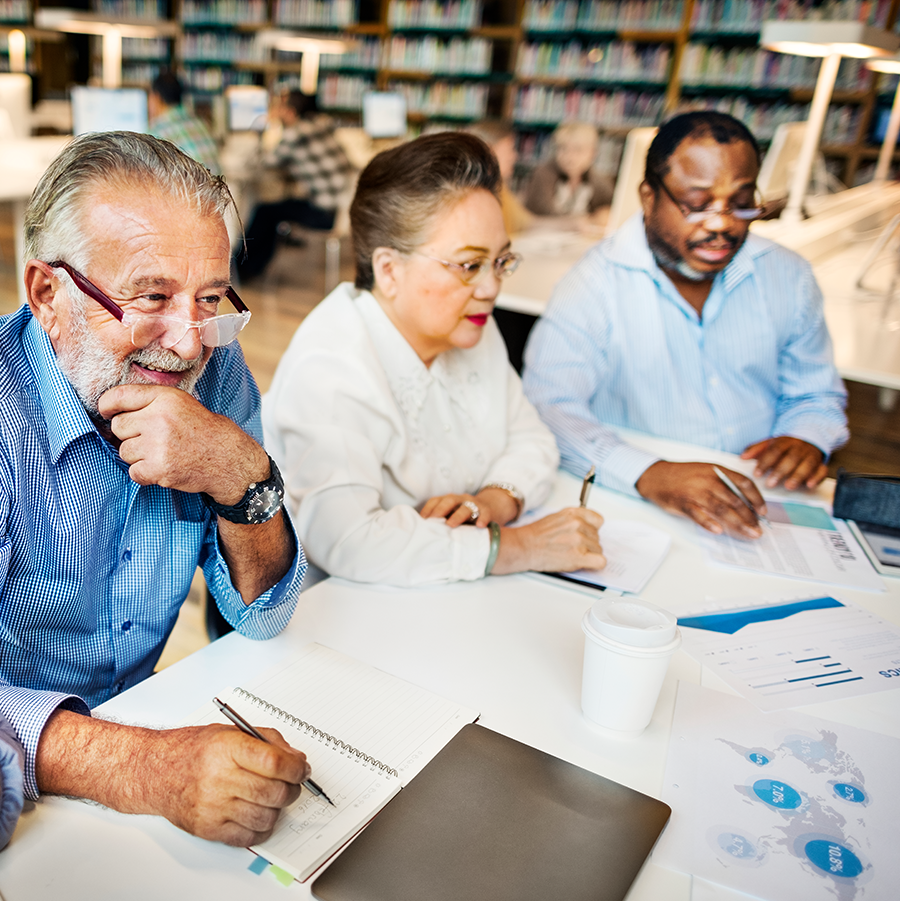 An older white man, Asian woman, and African American man sit at a table talking and looking at paperwork