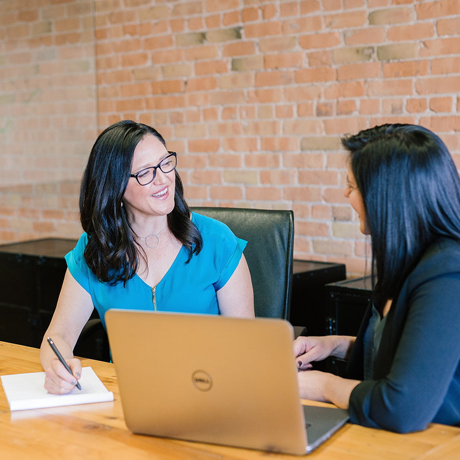 Two women sitting at a table talking. One is taking notes on a pad of paper and one has a laptop.